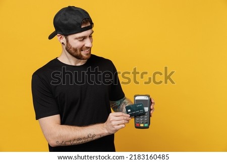 Young tattooed man 20s he wears casual black t-shirt cap hold wireless modern bank payment terminal process acquire credit card payments isolated on plain yellow wall background. Tattoo translate fun