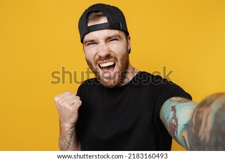 Close up young bearded tattooed man 20s he wears casual black t-shirt cap do selfie shot pov on mobile cell phone do winner gesture isolated on plain yellow wall background studio Tattoo translate fun