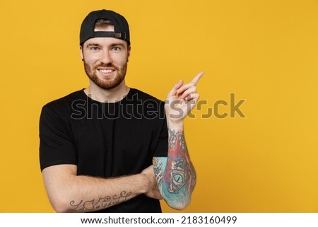Young happy bearded tattooed man 20s he wear casual black t-shirt cap indicate point index finger aside on workspace area mock up isolated on plain yellow wall background studio. Tattoo translate fun