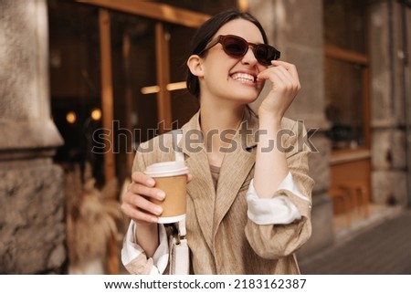 Joyful young caucasian woman enjoying her weekend drinking coffee outdoors. Brunette wears sunglasses, beige jacket. Good mood concept