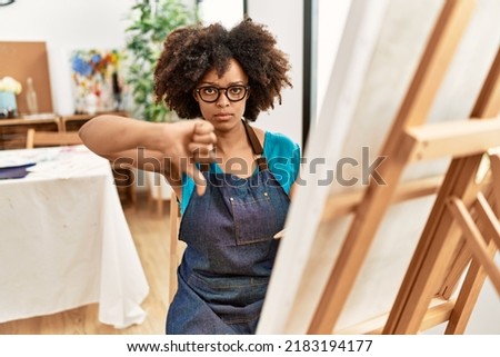 Beautiful african american woman with afro hair painting canvas at art studio with angry face, negative sign showing dislike with thumbs down, rejection concept 
