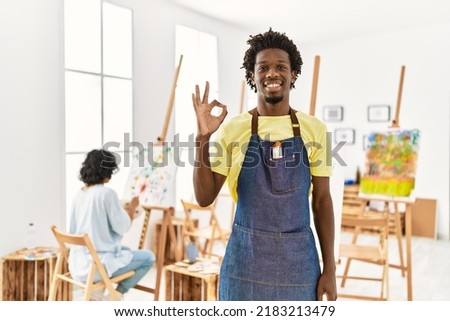 African young man standing at art studio smiling positive doing ok sign with hand and fingers. successful expression. 