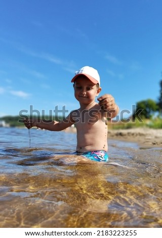 A little boy is sitting on the bank of a river in the water, playing with sand, with a white baseball cap on his head