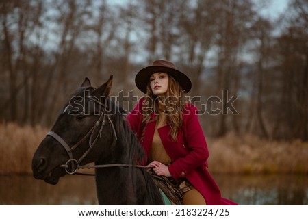 Elegant fashionable confident woman wearing trendy brown hat, classic red marsala color woolen coat posing, riding a horse in nature. Outdoor autumn fashion portrait. Copy, empty space for text
 Royalty-Free Stock Photo #2183224725