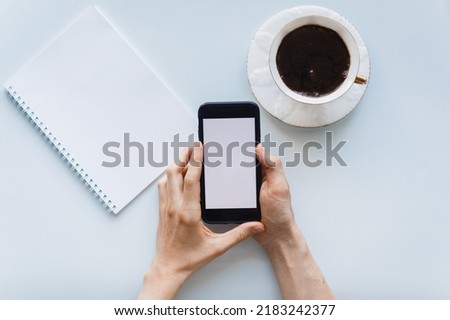 Top view composition on blue background. Girl holds mobile phone in hands, template for design. Cup of coffee, notebook for writing notes. Concept of online learning, scroll social networks and news.