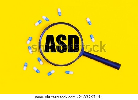 Pharmaceutical tablets and capsules near a magnifying glass with the text ASD Autism Spectrum Disorder on a yellow background. View from above.