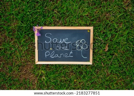 Black board with wooden edge and a pink flower with the message and a flower written with coloured chalks save the planet on a background of green grass in nature. 