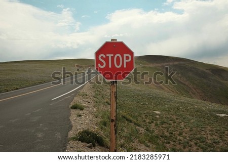 Red stop sign in center of photo on empty, deserted winding road in the green hills or mountains with cloudy blue sky in background
