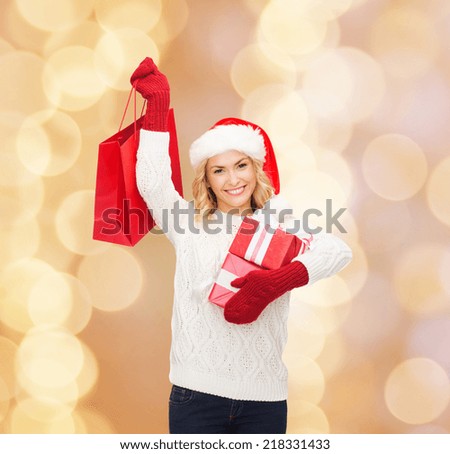 happiness, winter holidays, christmas and people concept - smiling young woman in santa helper hat with gifts and shopping bag over beige lights background