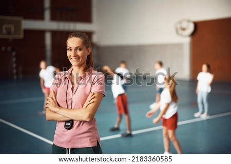 Confident female coach during physical education class at school gym looking at camera. Group of kids are exercising in the background. 