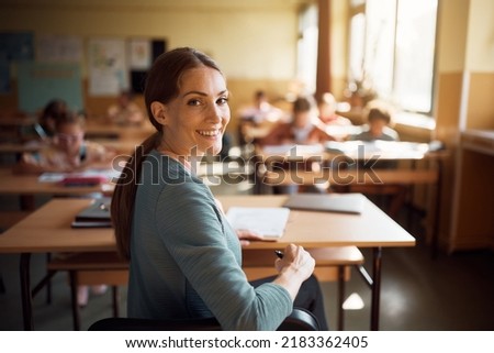 Happy elementary school teacher during a class looking at camera. Her students are leaning in the background.