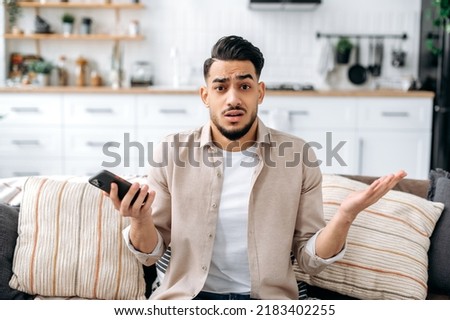 Confused puzzled indian or arabian guy in casual clothes, sits on a sofa in an interior living room, holds a smartphone in his hand, looks questioningly at the camera, spreading his arms around Royalty-Free Stock Photo #2183402255