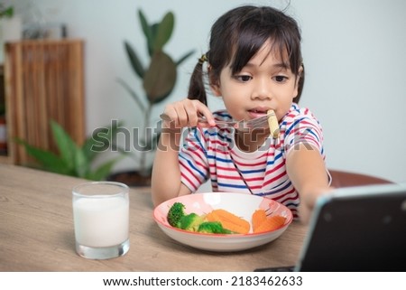 Adorable Asia child girl having lunch while watching a movie from the tablet. A little Asian child eating dinner and eyes are looking cartoon from Tablet. National Eating Disorders Awareness Week.