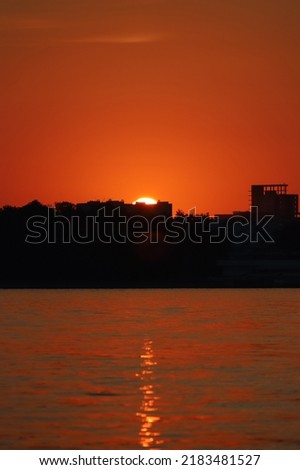 A bright setting sun sets behind the black silhouettes of buildings above the river against an orange red sky. Summer sunset background. Beautiful landscape, close up