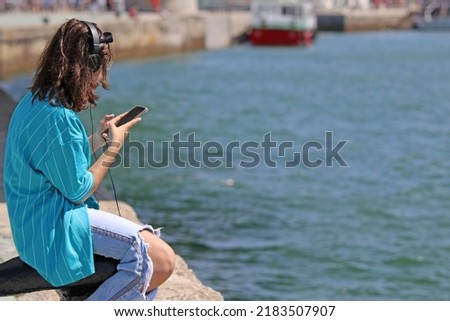 Young girl in casual clothes listening to music and looking at the mobile by the sea