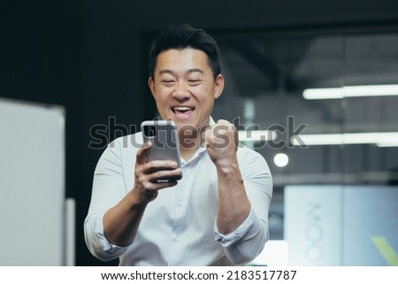 A break at work. A young Asian man holds a phone in his hands, plays games on the phone in the office at work, rejoices, shows with his hand yes,rejoices at the win, won a prize, rests, does not work. Royalty-Free Stock Photo #2183517787