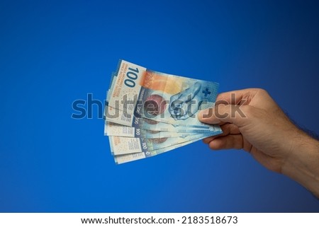 A spread of 100 Swiss Francs banknotes, held in hand by a Caucasian male. Close up studio shot, isolated on blue background