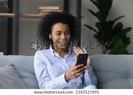 Smiling African American woman in wireless earphones waving hand at phone camera, holding smartphone, happy young female chatting with friends online by video call at home, blogger greeting viewers
