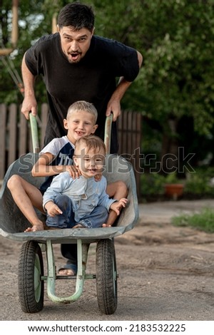 Dad has fun with the kids on the street on a summer day. The father drives the children in a wheelbarrow through the countryside. The concept of a happy childhood