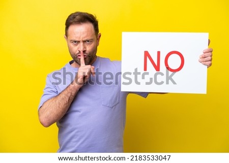 Middle age caucasian man isolated on yellow background holding a placard with text NO doing silence gesture