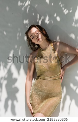 pretty young woman with closed eyes posing with hand on hip near white wall with shadows from leaves