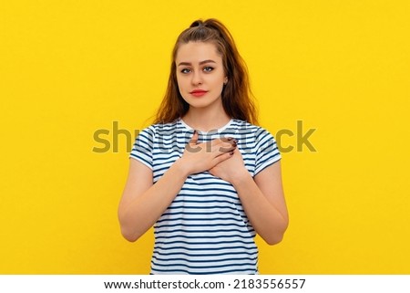 Portrait of peaceful, dear, good young woman holding hands on heart in appreciation, thank you gesture, being touched or flattered, standing in white-blue striped t shirt over yellow background