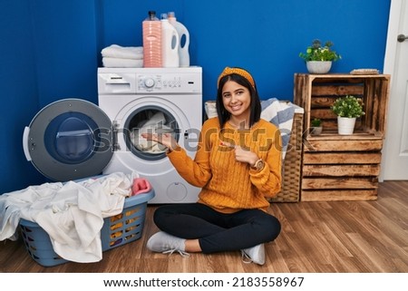 Young hispanic woman doing laundry amazed and smiling to the camera while presenting with hand and pointing with finger. 