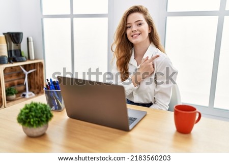 Young caucasian woman working at the office using computer laptop cheerful with a smile on face pointing with hand and finger up to the side with happy and natural expression 