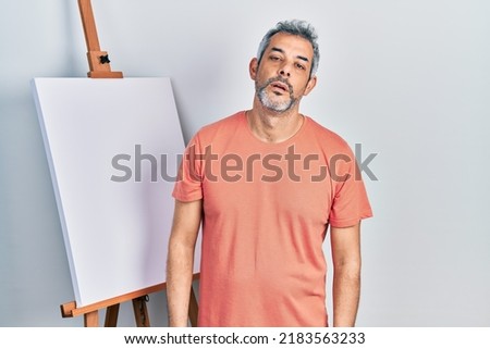 Handsome middle age man with grey hair standing by painter easel stand looking sleepy and tired, exhausted for fatigue and hangover, lazy eyes in the morning.  Royalty-Free Stock Photo #2183563233