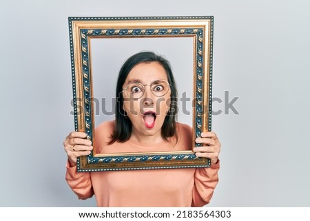 Middle age hispanic woman holding empty frame afraid and shocked with surprise and amazed expression, fear and excited face. 