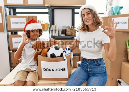 Young mother with little son wearing christmas hat at donations stand doing ok sign with fingers, smiling friendly gesturing excellent symbol 