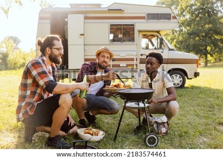 A group of friends spend time together in nature. Middle-aged men prepare a barbecue near an RV