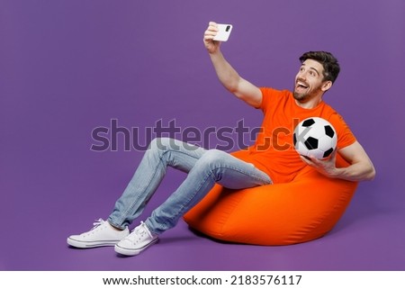 Full body young fan man he wear t-shirt cheer up support football team hold soccer ball watch tv live stream sit in bag chair do selfie shot mobile cell phone isolated on plain dark purple background
