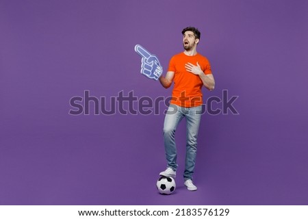 Full body young fan man he wear orange t-shirt cheer up support football sport team hold soccer ball # 1 foam glove finger up sing anthem watch tv live stream isolated on plain dark purple background Royalty-Free Stock Photo #2183576129