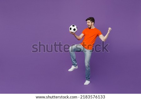 Full body size length sideways young fan man he wear orange t-shirt cheer up support football sport team hold juggling on knee soccer ball watch tv live stream isolated on plain dark purple background
