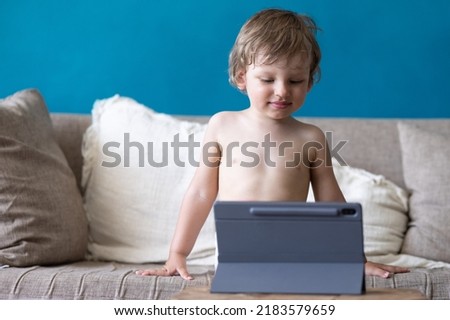 Cute toddler child watches cartoons or playing games using a tablet pc, sitting on sofa at home. Gadgets and modern entertainment for children. Video chat, video call.