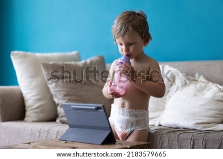 Cute toddler child watches cartoons or playing games using a tablet pc, sitting on sofa at home and drinking water. Gadgets and modern entertainment for children. Video chat, video call.