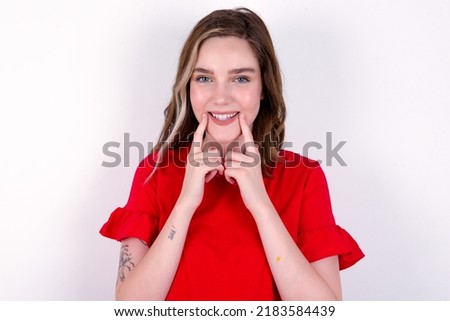 Happy young caucasian woman wearing red T-shirt over white background with toothy smile, keeps index fingers near mouth, fingers pointing and forcing cheerful smile