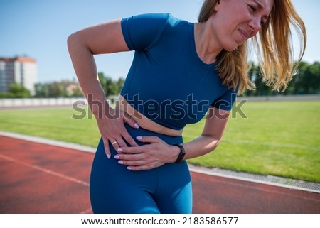 Girl in sportswear in stadium is exhausted and has pain during training and workout.Sports woman has a stomach ache. Outdoor sports and pain concept. Royalty-Free Stock Photo #2183586577