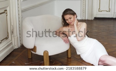 feminine photo shoot. an attractive woman in a white dress with open shoulders poses for a photographer on the floor by the chair. femininity. fashionable women's clothing store. Royalty-Free Stock Photo #2183588879