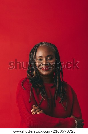 Portrait of a woman with dreadlocks looking at the camera while standing against a red background with her arms crossed. Mature black woman embracing her natural hair with confidence.