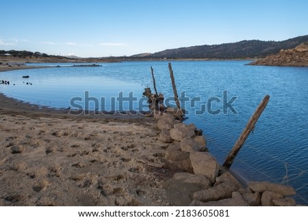 A submerged and weathered barbed wire fence enters the water of a lake. The rusty iron poles are worn and worm-eaten, the barbed wire is rusty.