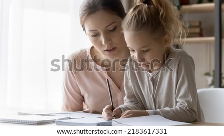 Small Caucasian girl child and young mother study together at home, prepare homework write in notebook. Loving caring mom help small daughter with hometask handwrite take notes. Education concept. Royalty-Free Stock Photo #2183617331