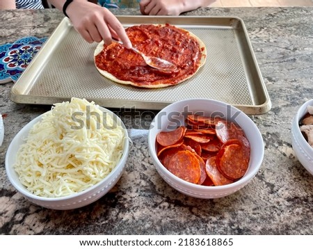 The top down, close up image of a child making a homemade pizza on a cauliflower crust. She's spooning sauce and has bowls filled with toppings.