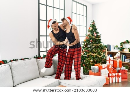 Two hispanic men couple dancing and standing on sofa by christmas tree at home