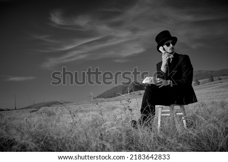 A serious man in a black suit, top hat and black sunglasses sits on a stool in a field and calls on the phone. Black-and-white photo. Surrealism. Copy space.