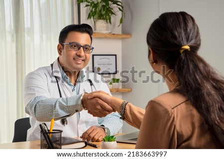 An Indian young man or male smiling physician or doctor sitting in a modern clinic wearing a stethoscope and apron shaking hand with a happy female patient. Medical, medicine, and healthcare concept Royalty-Free Stock Photo #2183663997