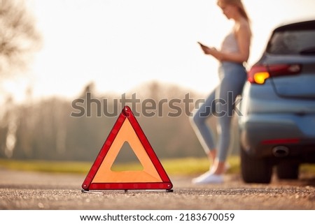 Hazard Warning Triangle Sign For Car Breakdown On Road With Woman Calling For Help In Background Royalty-Free Stock Photo #2183670059