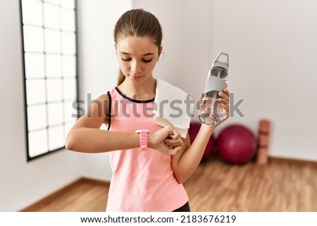 Young brunette teenager wearing sportswear holding water bottle checking the time on wrist watch, relaxed and confident 