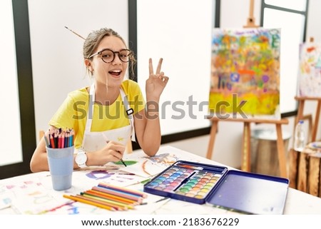 Young brunette teenager at art studio smiling with happy face winking at the camera doing victory sign with fingers. number two. 